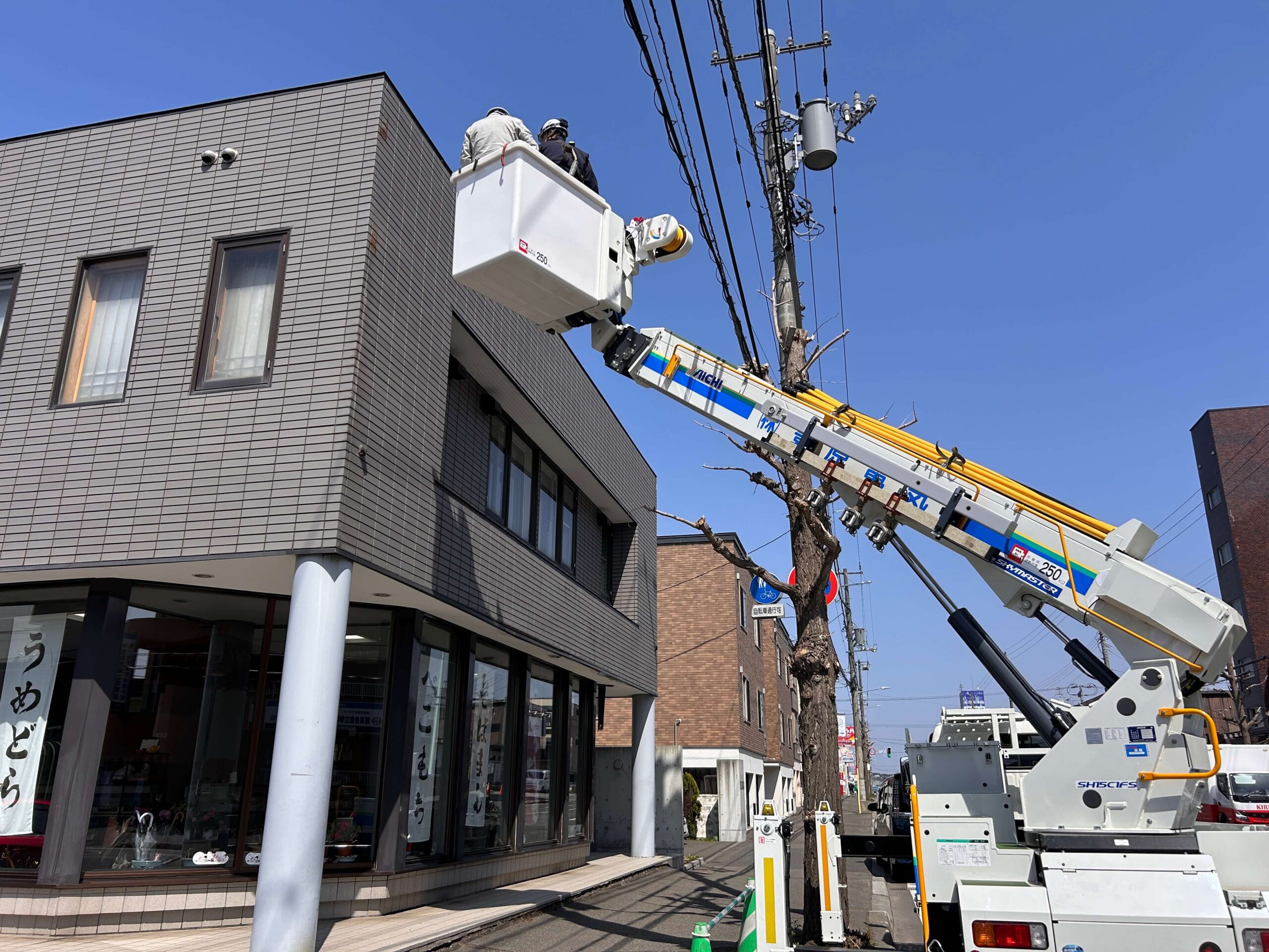 作業中（高所）：高所作業車のバスケット内から看板の配線処理・ビス取り外し作業を行う作業員。