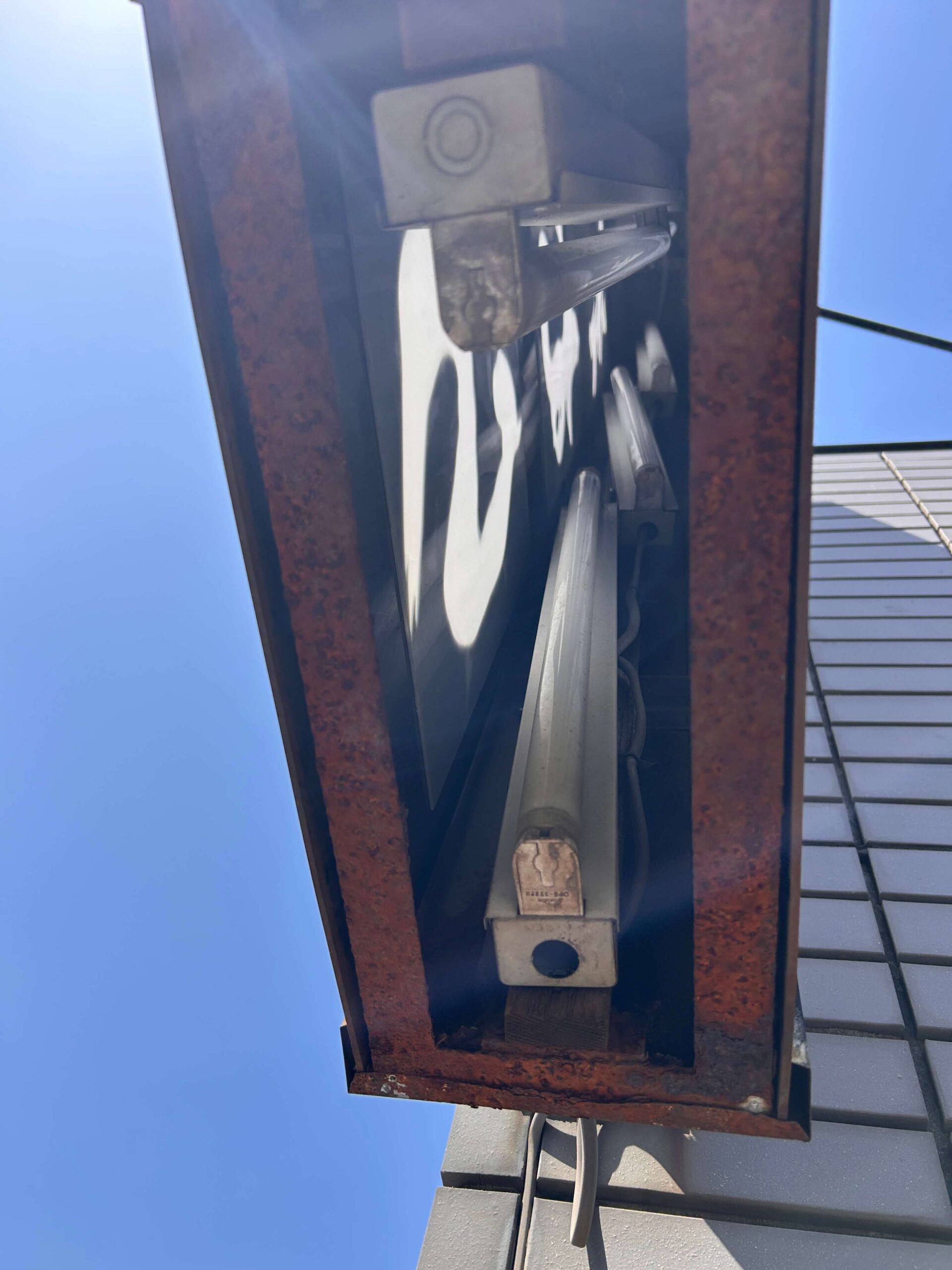 施工前（内部確認）：看板底面から内部を覗いた状態。蛍光管・反射板・金属フレームが露出しており、鉄部の錆が著しく進行している。