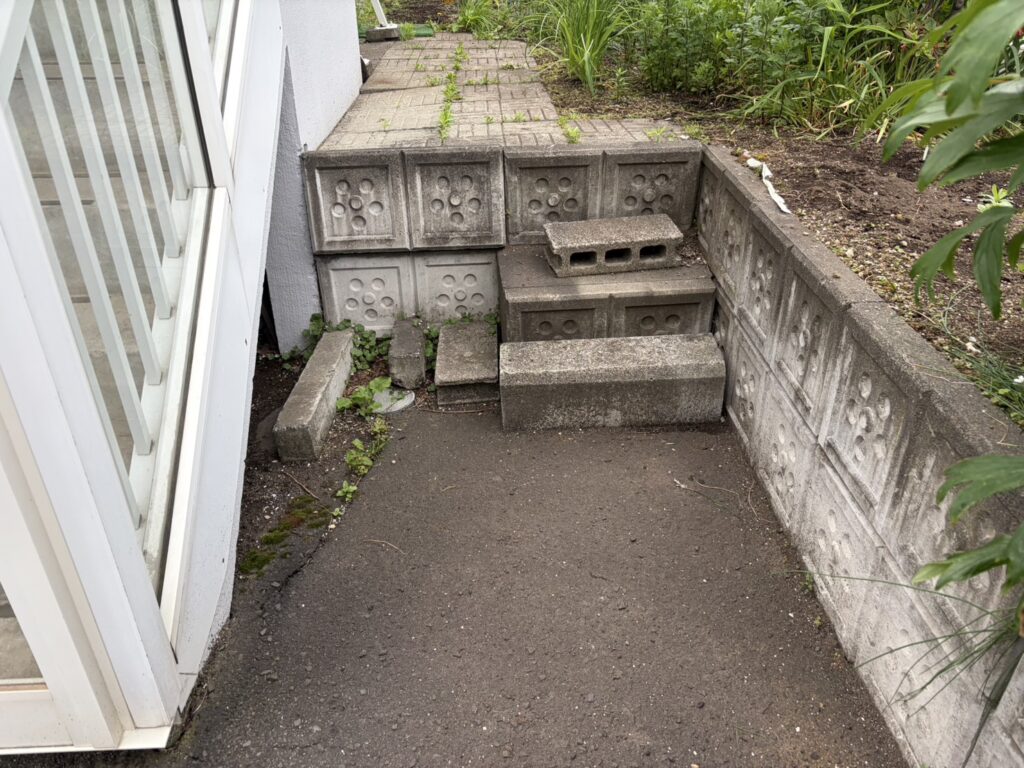 犬走り 防草シート施工前の状態。建物外壁ぎわにブロック塀と雑草が茂り、砂利も散乱している