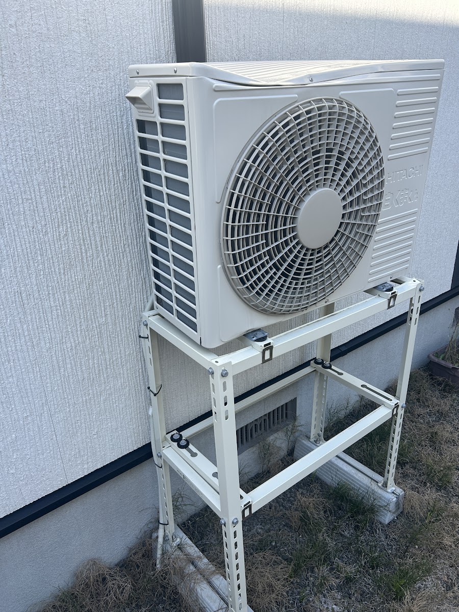 交換前の室外機と歪んだ白い架台。屋根はなく壁際に設置されている