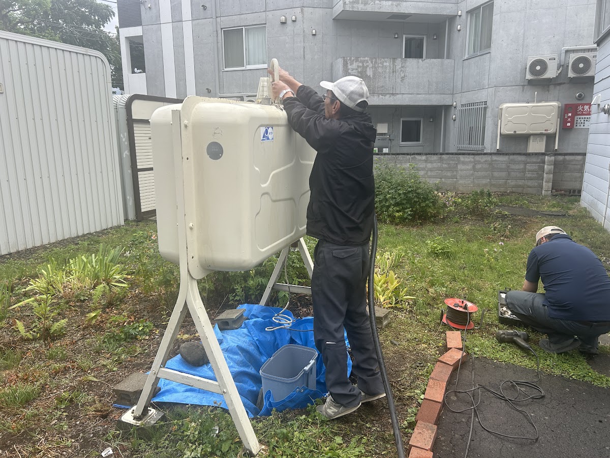 タンク上部で作業する作業員と、奥で工具を準備するもう一人の作業員。タンクの下にブルーシートとバケツがある